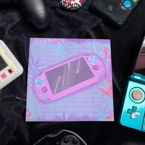Könnte beinhalten: Ein quadratischer Druck mit einer rosa Handheld-Spielkonsole mit blauen Akzenten, vor einem pastellfarbenen Hintergrund mit schneeflockenartigen Mustern. Um den Druck herum befinden sich verschiedene Gamecontroller und Konsolen, darunter ein schwarzer Controller und ein türkisfarbenes Handheld-Gerät.
