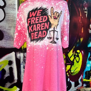 May include: Pink t-shirt with a bleached design. The shirt features the text "WE FREED KAREN READ" in red and black, along with a hand gesture and scales of justice graphic. The shirt has a round neck and three-quarter sleeves.