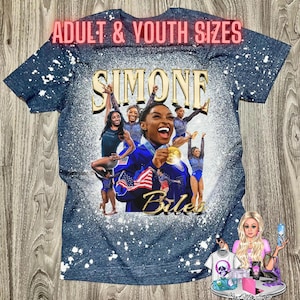 May include: A blue t-shirt with a white splatter paint design. The shirt features a photo of Simone Biles with the text "Simone Biles" and "Adult & Youth Sizes".