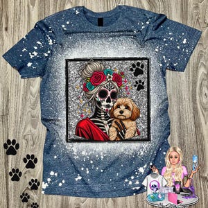 May include: Blue t-shirt with a graphic of a sugar skull woman holding a small dog. The woman has a floral crown and the dog is brown. The shirt has a bleached, speckled design and paw prints.