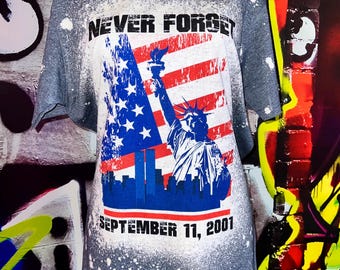 May include: A gray t-shirt with a white bleach splattered design. The design features a blue and red American flag with the Statue of Liberty holding a torch. The text "NEVER FORGET SEPTEMBER 11, 2001" is printed below the image.