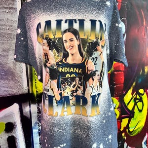 Puede incluir: Una camiseta gris con un dise&ntilde;o salpicado de lej&iacute;a que presenta a una jugadora de baloncesto con el nombre "CAITLIN CLARK" en letras amarillas y negras. La jugadora lleva una camiseta azul y amarilla con la palabra "INDIANA" en ella.