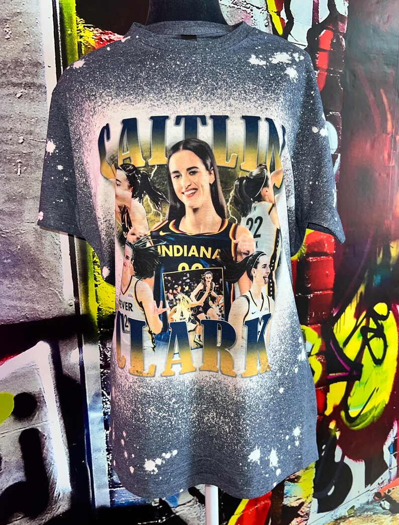Puede incluir: Una camiseta gris con un dise&ntilde;o salpicado de lej&iacute;a que presenta a una jugadora de baloncesto con el nombre "CAITLIN CLARK" en letras amarillas y negras. La jugadora lleva una camiseta azul y amarilla con la palabra "INDIANA" en ella.