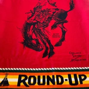 Puede incluir: Manta roja Pendleton con una ilustraci&oacute;n negra de un jinete de rodeo sobre un caballo salvaje. La manta tiene las palabras "PENDLETON" y "ROUND-UP" en negro, con rayas coloridas y dise&ntilde;os de tipi.