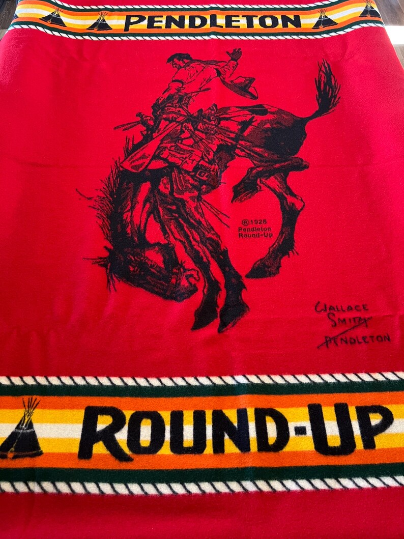 Puede incluir: Manta roja Pendleton con una ilustraci&oacute;n negra de un jinete de rodeo sobre un caballo salvaje. La manta tiene las palabras "PENDLETON" y "ROUND-UP" en negro, con rayas coloridas y dise&ntilde;os de tipi.