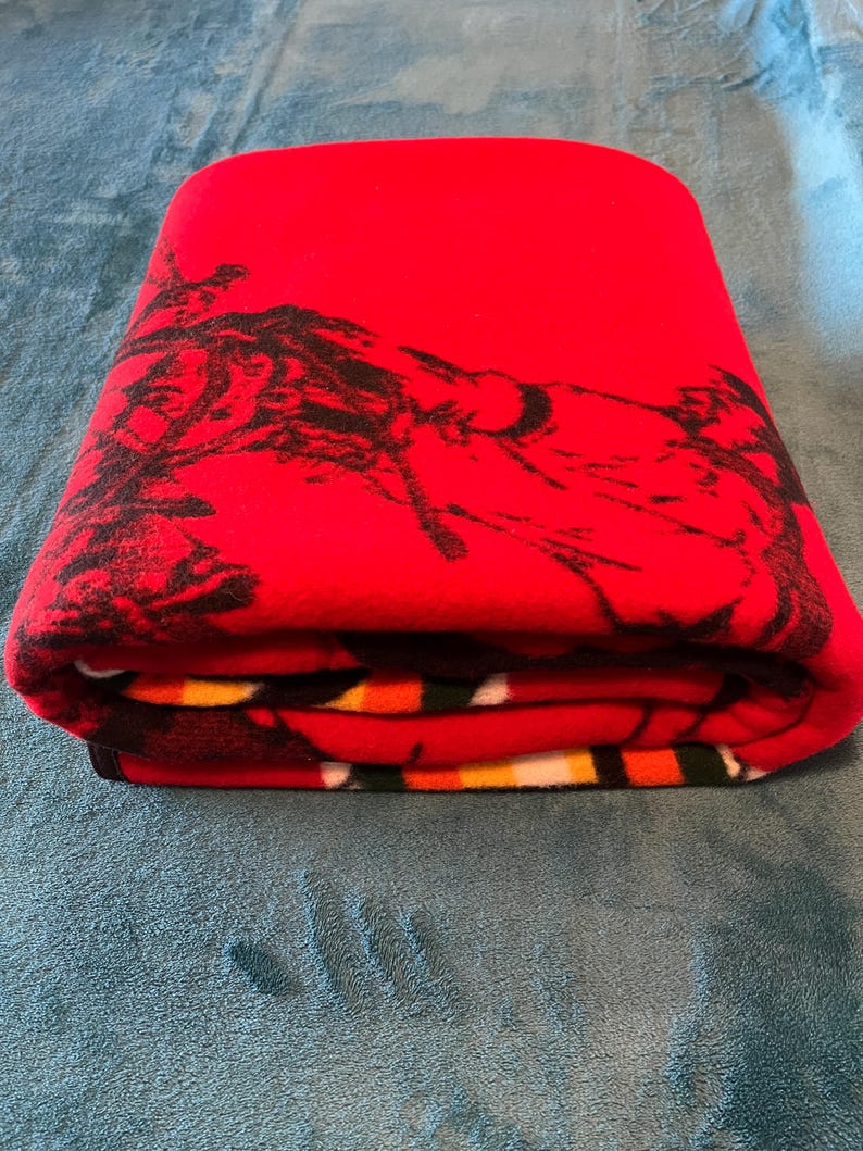 Puede incluir: Manta roja doblada con un dise&ntilde;o gr&aacute;fico negro. La manta est&aacute; doblada cuidadosamente, mostrando el dise&ntilde;o en la capa superior. La manta est&aacute; hecha de un material suave y est&aacute; doblada sobre una superficie azul.