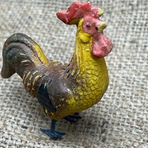 K&ouml;nnte beinhalten: Eine kleine, detaillierte Figur eines Hahns. Der Hahn hat einen roten Kamm und Kehllappen, einen gelben K&ouml;rper und braune und schwarze Federn. Die Figur steht auf einer strukturierten Oberfl&auml;che.