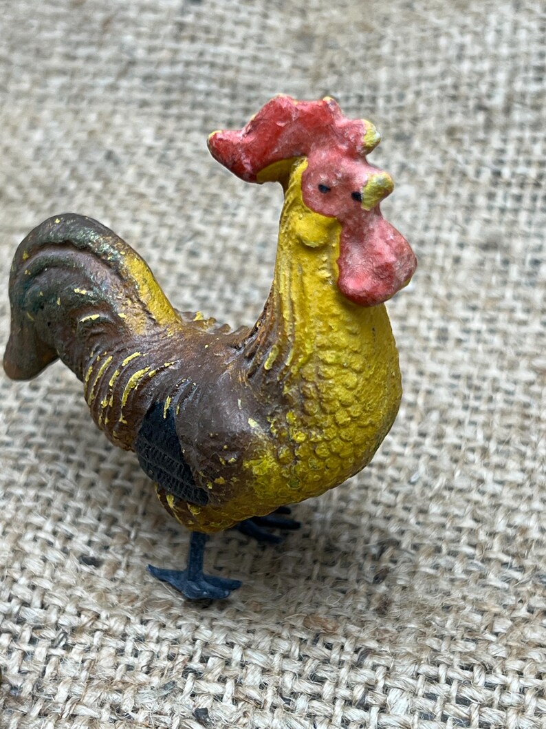 K&ouml;nnte beinhalten: Eine kleine, detaillierte Figur eines Hahns. Der Hahn hat einen roten Kamm und Kehllappen, einen gelben K&ouml;rper und braune und schwarze Federn. Die Figur steht auf einer strukturierten Oberfl&auml;che.