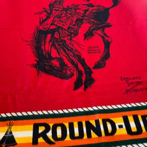 Puede incluir: Una manta roja Pendleton Round-Up con una ilustraci&oacute;n en negro de un vaquero de rodeo montando un bronco. La manta tiene un borde con las palabras "ROUND-UP" en letras negras y un gr&aacute;fico de tipi. La manta est&aacute; hecha de un material suave.