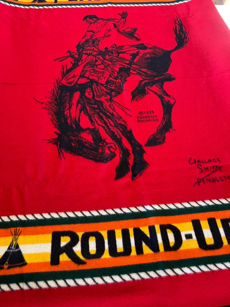 Puede incluir: Una manta roja Pendleton Round-Up con una ilustraci&oacute;n en negro de un vaquero de rodeo montando un bronco. La manta tiene un borde con las palabras "ROUND-UP" en letras negras y un gr&aacute;fico de tipi. La manta est&aacute; hecha de un material suave.