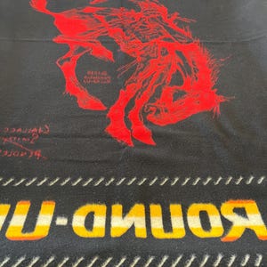 Puede incluir: Manta negra con un gr&aacute;fico rojo de un vaquero en un caballo salvaje. La manta tiene la palabra "ROUND-UP" en amarillo y naranja, y la palabra "PENDLETON" en la parte superior. La manta tiene un tema occidental.