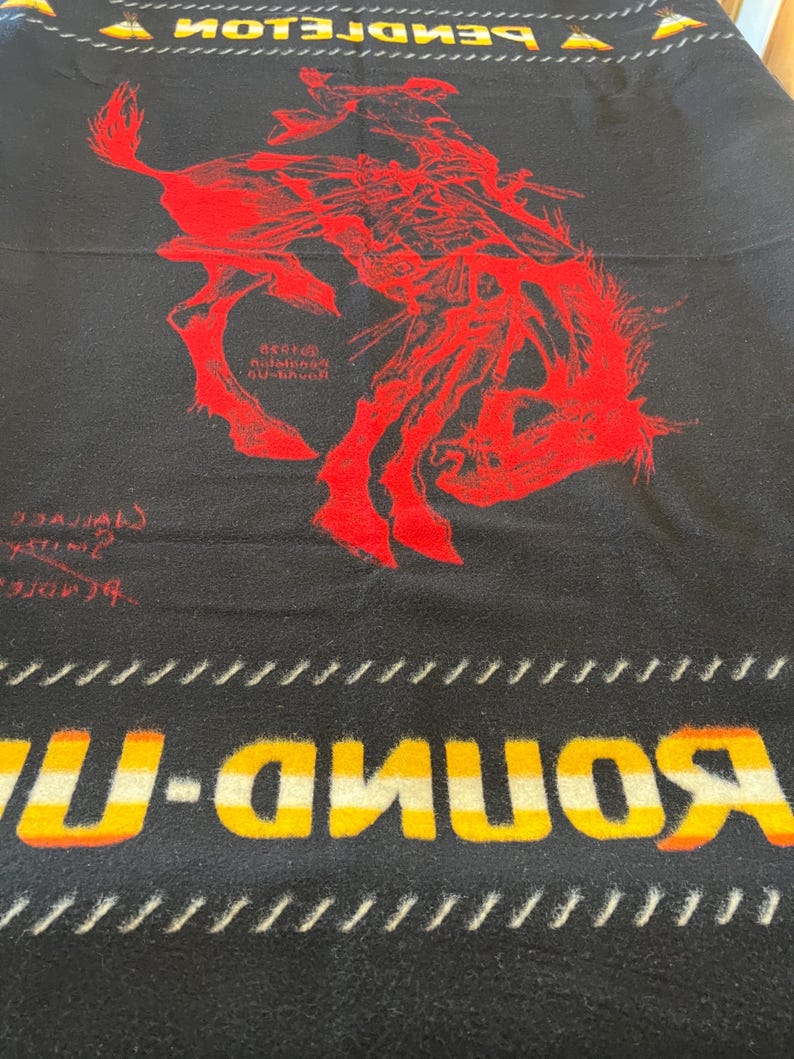 Puede incluir: Manta negra con un gr&aacute;fico rojo de un vaquero en un caballo salvaje. La manta tiene la palabra "ROUND-UP" en amarillo y naranja, y la palabra "PENDLETON" en la parte superior. La manta tiene un tema occidental.