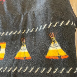 Puede incluir: Manta negra con un dise&ntilde;o nativo americano. La manta presenta im&aacute;genes repetidas de tipis en amarillo, naranja y blanco. Las costuras blancas bordean los bordes de la manta.