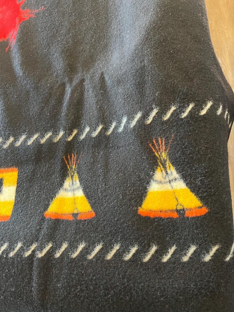 Puede incluir: Manta negra con un dise&ntilde;o nativo americano. La manta presenta im&aacute;genes repetidas de tipis en amarillo, naranja y blanco. Las costuras blancas bordean los bordes de la manta.
