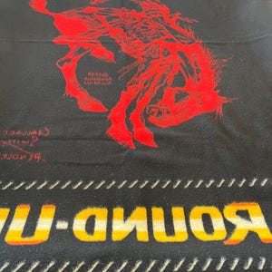 Puede incluir: Manta negra con una ilustraci&oacute;n roja de un jinete de rodeo sobre un bronco. La manta tiene las palabras "ROUND-UP" en amarillo y blanco, y "PENDLETON" en amarillo. La manta tiene un borde blanco y negro.