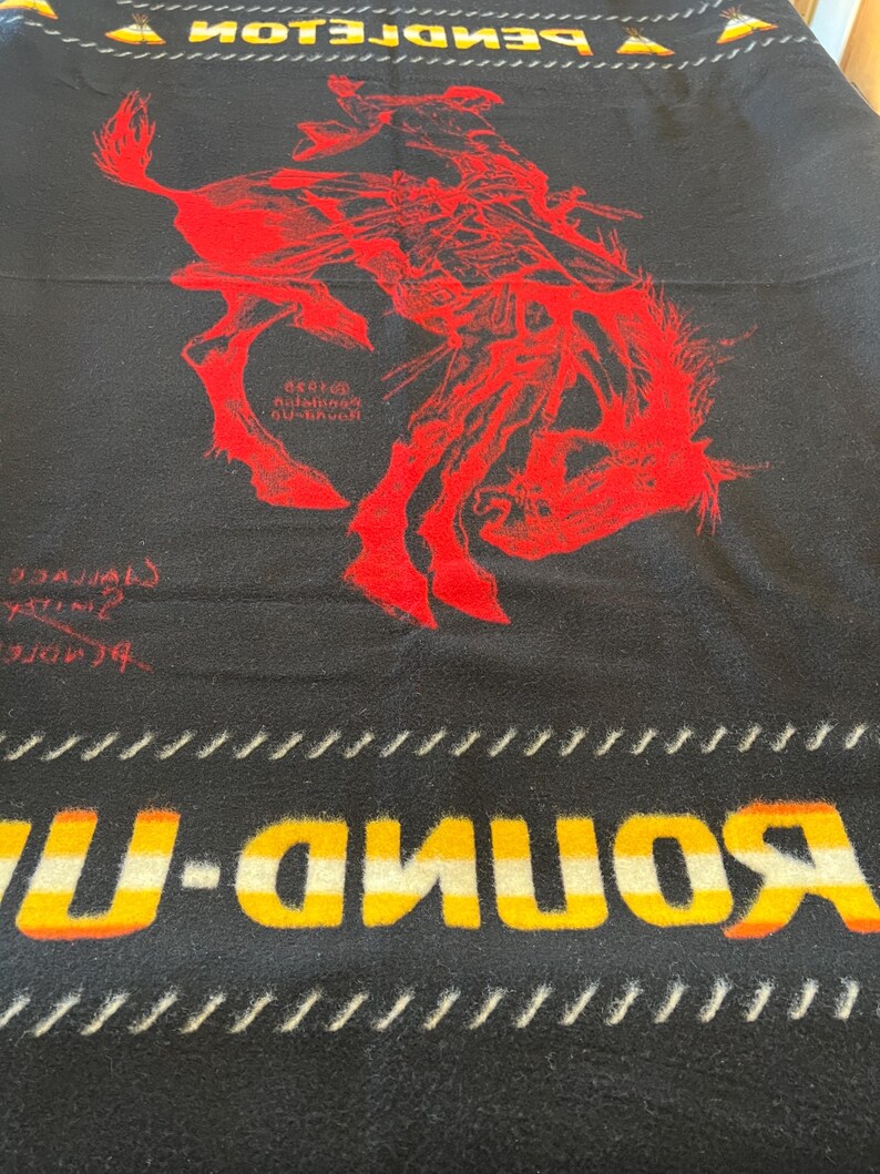 Puede incluir: Manta negra con una ilustraci&oacute;n roja de un jinete de rodeo sobre un bronco. La manta tiene las palabras "ROUND-UP" en amarillo y blanco, y "PENDLETON" en amarillo. La manta tiene un borde blanco y negro.