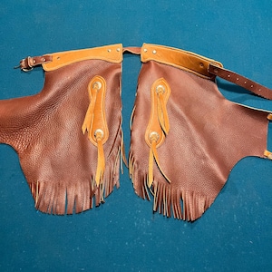 May include: Brown leather chaps with tan leather accents. The chaps feature decorative silver-toned hardware, fringe along the bottom, and adjustable straps. These chaps are designed for riding.