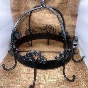 May include: A black metal hanging pot rack with decorative scrollwork and six hooks. The rack has a circular design with a top loop for hanging. The metal has a textured finish, and the rack is displayed against a burlap background.