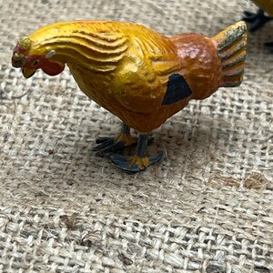 K&ouml;nnte beinhalten: Kleine, dekorative H&uuml;hnerfigur mit gelb-orangefarbenem K&ouml;rper, schwarzem Fleck und rotem Kamm. Die Figur hat detaillierte Merkmale und steht auf winzigen F&uuml;&szlig;en. Der Hintergrund ist ein strukturiertes, gewebtes Gewebe.