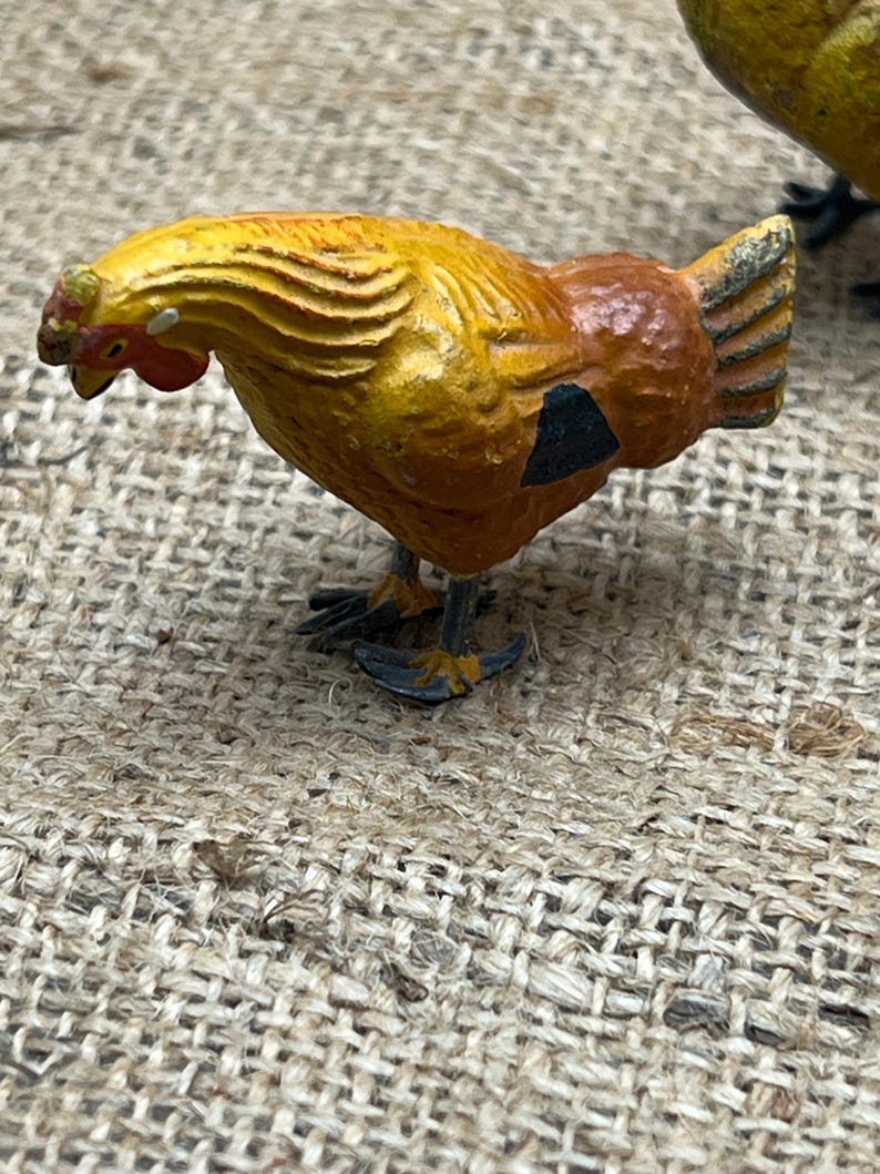 K&ouml;nnte beinhalten: Kleine, dekorative H&uuml;hnerfigur mit gelb-orangefarbenem K&ouml;rper, schwarzem Fleck und rotem Kamm. Die Figur hat detaillierte Merkmale und steht auf winzigen F&uuml;&szlig;en. Der Hintergrund ist ein strukturiertes, gewebtes Gewebe.