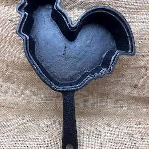 May include: Black cast iron skillet in the shape of a rooster. The skillet has a handle with a hole for hanging. The rooster-shaped pan is designed for cooking and serving food. The pan is sitting on a burlap surface.