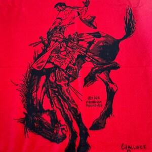 Puede incluir: Textil rojo con una ilustraci&oacute;n negra de un vaquero montando un bronco. El borde superior muestra la palabra "PENDLETON" sobre un dise&ntilde;o de inspiraci&oacute;n nativa americana. Tambi&eacute;n se ve el texto "@1925 Pendleton Round-Up" y la firma "Wallace Smith Pendleton".