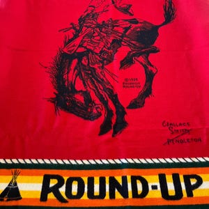 May include: A red Pendleton blanket featuring a black illustration of a cowboy riding a bucking bronco. The blanket has the words "PENDLETON" and "ROUND-UP" in white and black, with a colorful striped border. The blanket also has the text "©1926 Pendleton Round-Up" and the signature of Wallace Smith.