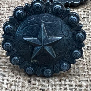 May include: Close-up of a dark metal concho with a star in the center and a decorative floral pattern. The concho is surrounded by small, circular embellishments. The background is a textured, tan fabric.