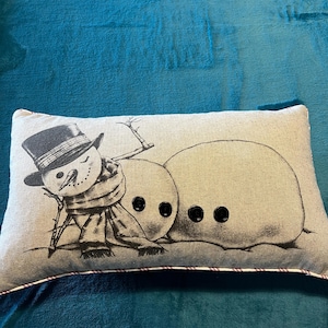 May include: Rectangular pillow with a whimsical snowman design. The pillow features a black and white illustration of a snowman wearing a top hat and scarf, leaning against two snowballs with button eyes. The pillow has a neutral background and a red and white trim.