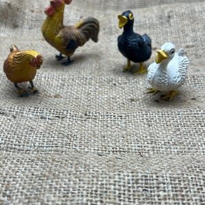 K&ouml;nnte beinhalten: Vier Miniatur-Bauernhoftierfiguren: ein gelb-brauner Hahn, eine kleine gelbe Henne, eine schwarze Ente und eine wei&szlig;e Ente mit gelbem Schnabel. Die Figuren sind vor einem Jutegewebe-Hintergrund platziert.