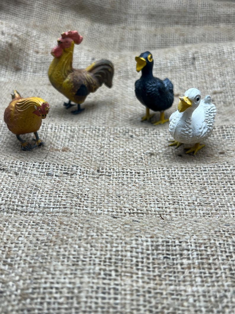 K&ouml;nnte beinhalten: Vier Miniatur-Bauernhoftierfiguren: ein gelb-brauner Hahn, eine kleine gelbe Henne, eine schwarze Ente und eine wei&szlig;e Ente mit gelbem Schnabel. Die Figuren sind vor einem Jutegewebe-Hintergrund platziert.