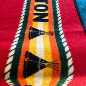 Puede incluir: Una colorida manta con la palabra "OLETON" en letras negras grandes en el centro. La manta presenta un fondo rojo con un dise&ntilde;o central de rayas naranjas, amarillas y blancas, y un borde verde. Tambi&eacute;n se ven dos dise&ntilde;os gr&aacute;ficos negros y amarillos.