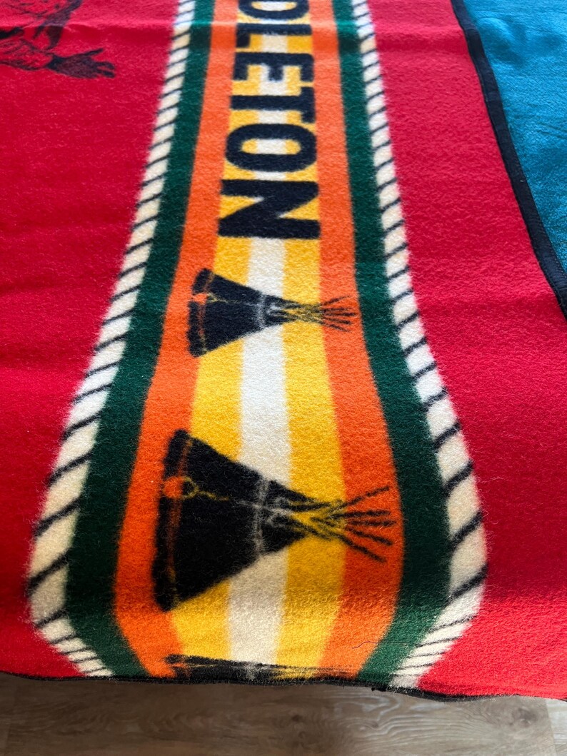 Puede incluir: Una colorida manta con la palabra "OLETON" en letras negras grandes en el centro. La manta presenta un fondo rojo con un dise&ntilde;o central de rayas naranjas, amarillas y blancas, y un borde verde. Tambi&eacute;n se ven dos dise&ntilde;os gr&aacute;ficos negros y amarillos.
