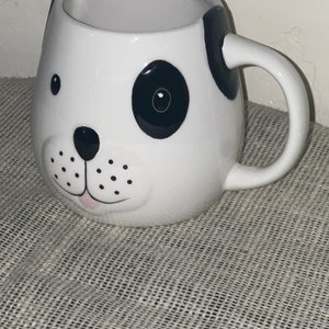 May include: White ceramic mug shaped like a dog's face with black and white markings. The mug has a black nose, black eyes, and a pink tongue.