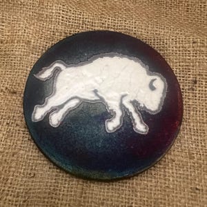 May include: A round, dark blue coaster with a white buffalo design. The buffalo is in a running pose, with a detailed outline. The coaster has a textured, iridescent finish with hints of red and green.
