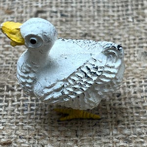 K&ouml;nnte beinhalten: Kleine, wei&szlig;e Entenfigur mit gelbem Schnabel und F&uuml;&szlig;en. Die Ente hat schwarze Akzente auf ihren Fl&uuml;geln und eine strukturierte Oberfl&auml;che, die ihr ein rustikales Aussehen verleiht. Die Figur steht vor einem neutralen, strukturierten Hintergrund.
