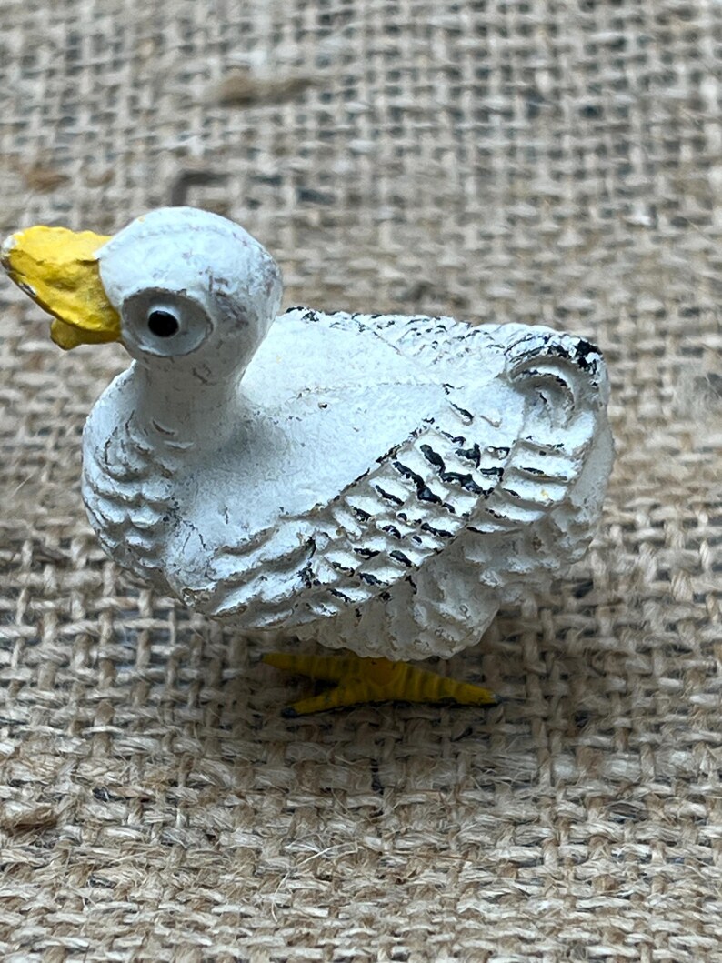 K&ouml;nnte beinhalten: Kleine, wei&szlig;e Entenfigur mit gelbem Schnabel und F&uuml;&szlig;en. Die Ente hat schwarze Akzente auf ihren Fl&uuml;geln und eine strukturierte Oberfl&auml;che, die ihr ein rustikales Aussehen verleiht. Die Figur steht vor einem neutralen, strukturierten Hintergrund.