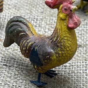 K&ouml;nnte beinhalten: Miniatur-Hahnfigur mit gelbem K&ouml;rper, braunen Fl&uuml;geln und rotem Kamm und Kehllappen. Die detaillierte Skulptur hat schwarze F&uuml;&szlig;e und ist auf einer strukturierten Oberfl&auml;che platziert, die ihren Vintage-Charme zeigt.