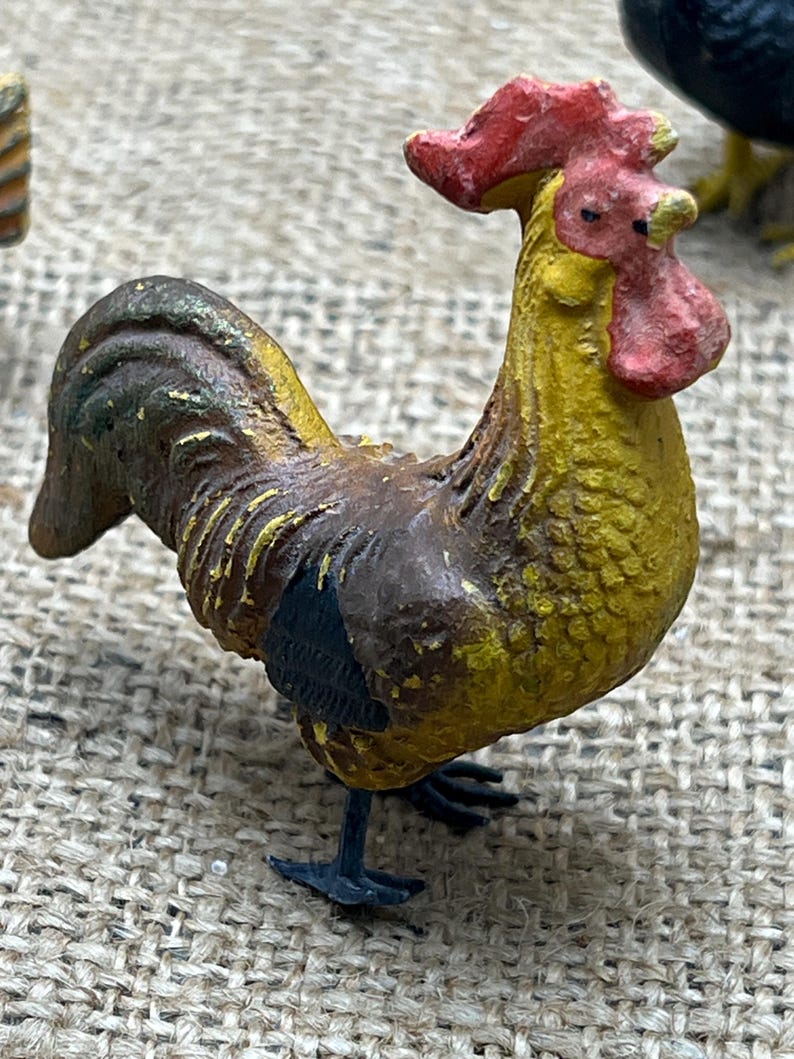 K&ouml;nnte beinhalten: Miniatur-Hahnfigur mit gelbem K&ouml;rper, braunen Fl&uuml;geln und rotem Kamm und Kehllappen. Die detaillierte Skulptur hat schwarze F&uuml;&szlig;e und ist auf einer strukturierten Oberfl&auml;che platziert, die ihren Vintage-Charme zeigt.