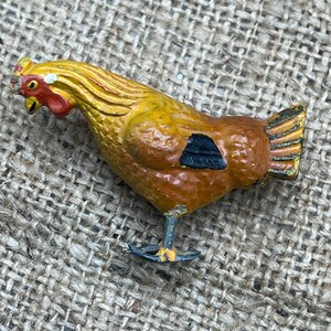 K&ouml;nnte beinhalten: Eine bemalte Metall-H&uuml;hnerfigur. Das Huhn hat einen gelben Kopf und Hals, einen braunen K&ouml;rper mit einer schwarzen Markierung und einen goldenen Schwanz. Die Figur steht auf zwei d&uuml;nnen Beinen.