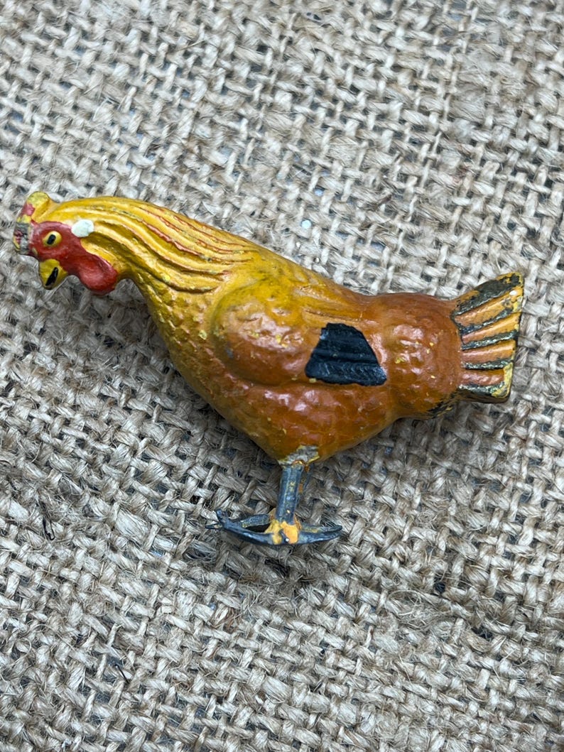 K&ouml;nnte beinhalten: Eine Vintage-H&uuml;hnerfigur aus bemaltem Metall. Das Huhn ist haupts&auml;chlich gelb und braun mit rotem Kamm und Kehllappen. Es hat eine schwarze Markierung an der Seite und steht auf einem kleinen, blauen Sockel. Die Figur befindet sich vor einem strukturierten, beigefarbenen Hintergrund.