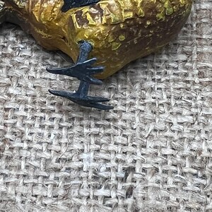 K&ouml;nnte beinhalten: Nahaufnahme einer dekorativen Hahnenfigur. Der Hahn ist in Gelb-, Braun- und Schwarzt&ouml;nen bemalt. Die Metallf&uuml;&szlig;e der Figur sind sichtbar, vor einem Hintergrund aus strukturiertem, gewebtem Stoff.