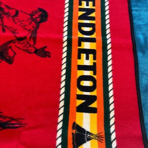 Puede incluir: Manta roja con una ilustraci&oacute;n negra de una persona corriendo. La manta tiene una franja vertical con la palabra "PENDLETON" en letras negras. La franja tiene detalles en verde, naranja, amarillo y blanco.