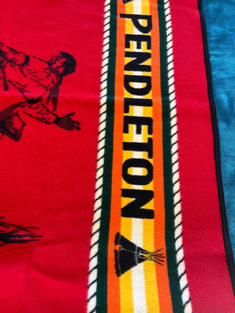 Puede incluir: Manta roja con una ilustraci&oacute;n negra de una persona corriendo. La manta tiene una franja vertical con la palabra "PENDLETON" en letras negras. La franja tiene detalles en verde, naranja, amarillo y blanco.