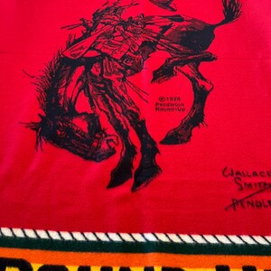 Puede incluir: Una manta roja de Pendleton con una ilustraci&oacute;n en negro de un vaquero montando un caballo salvaje. La manta tiene el texto "PENDLETON" en la parte superior y "ROUND-UP" en la inferior, con el a&ntilde;o "1926" visible.