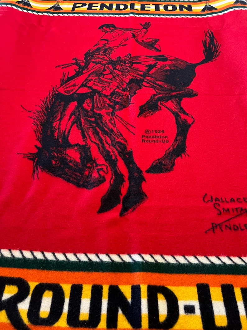 Puede incluir: Una manta roja de Pendleton con una ilustraci&oacute;n en negro de un vaquero montando un caballo salvaje. La manta tiene el texto "PENDLETON" en la parte superior y "ROUND-UP" en la inferior, con el a&ntilde;o "1926" visible.