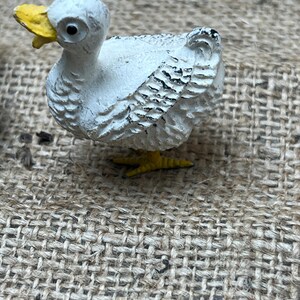 K&ouml;nnte beinhalten: Kleine, wei&szlig;e Entenfigur mit gelbem Schnabel und F&uuml;&szlig;en. Die Ente hat schwarze Details auf ihren Fl&uuml;geln und ist vor einem strukturierten, neutral get&ouml;nten Hintergrund platziert. Die Figur ist ein dekorativer Gegenstand.
