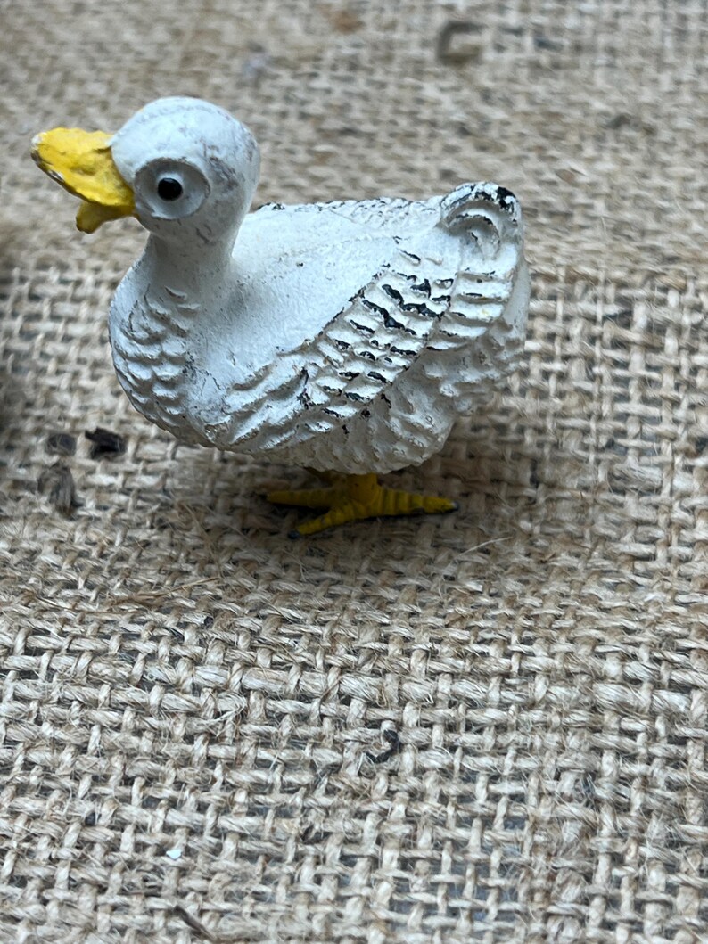 K&ouml;nnte beinhalten: Kleine, wei&szlig;e Entenfigur mit gelbem Schnabel und F&uuml;&szlig;en. Die Ente hat schwarze Details auf ihren Fl&uuml;geln und ist vor einem strukturierten, neutral get&ouml;nten Hintergrund platziert. Die Figur ist ein dekorativer Gegenstand.