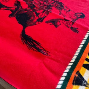 Puede incluir: Una manta roja con un gr&aacute;fico negro de una persona montando a caballo. La manta tiene un borde estampado con rayas verdes, blancas, amarillas y naranjas. La manta est&aacute; doblada.