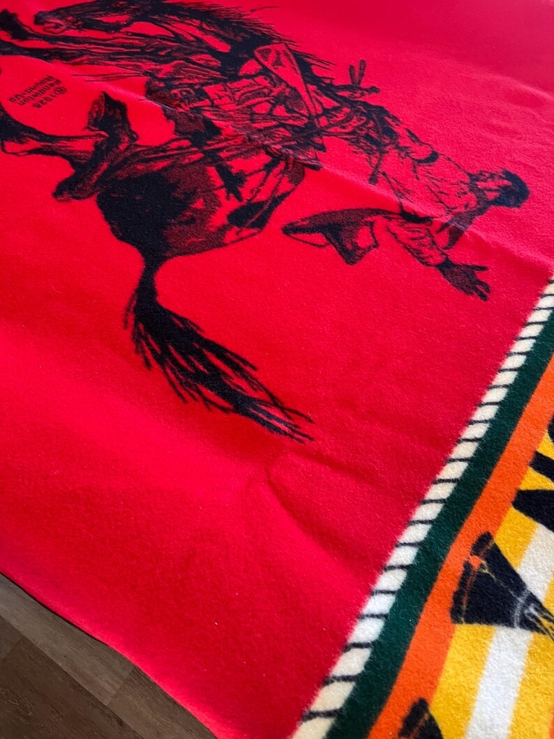 Puede incluir: Una manta roja con un gr&aacute;fico negro de una persona montando a caballo. La manta tiene un borde estampado con rayas verdes, blancas, amarillas y naranjas. La manta est&aacute; doblada.